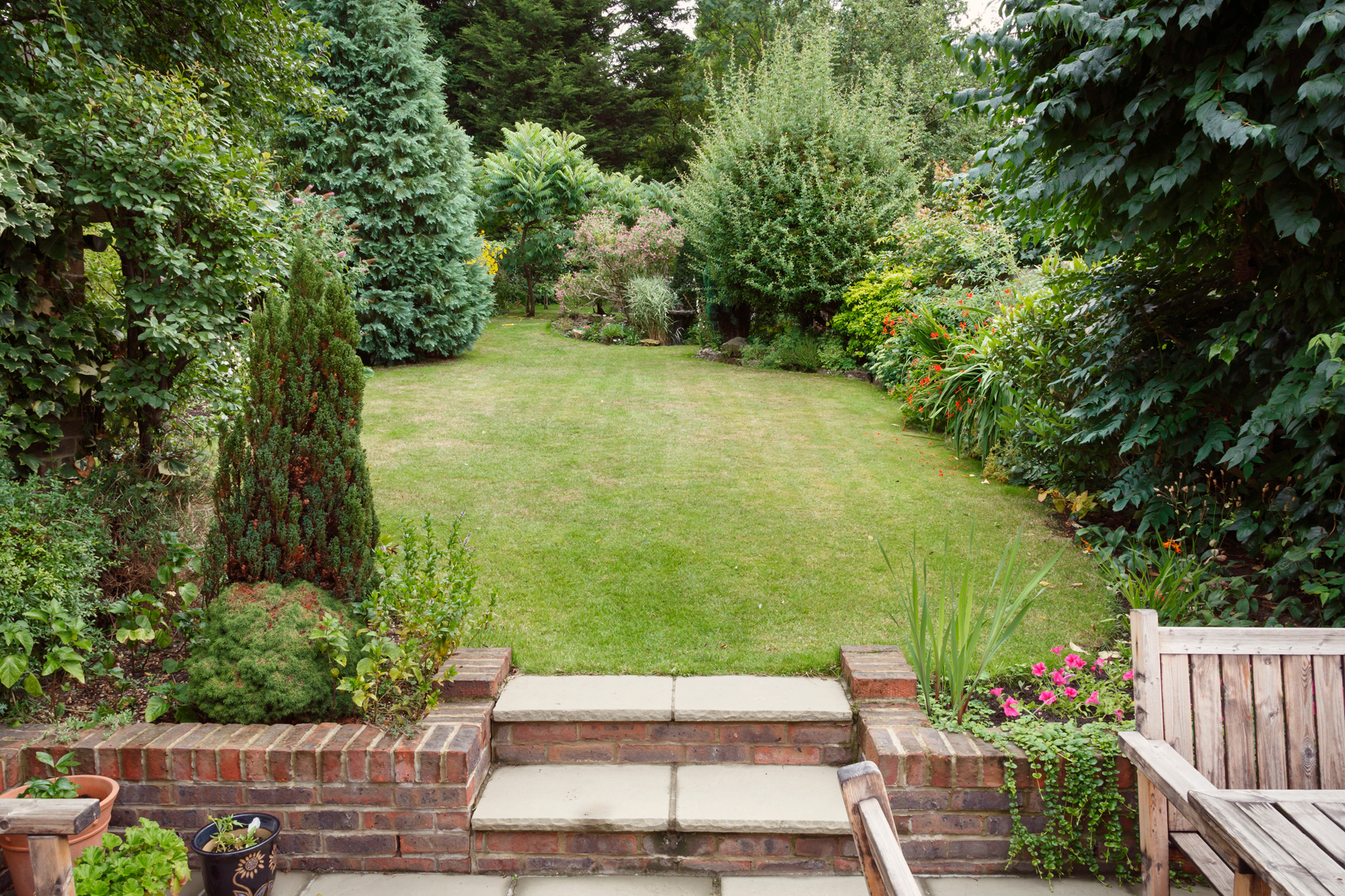 Kleingarten mit Terrasse, Gartenmöbeln und Pflanzenkübeln vorne, dahinter Rasenfläche mit kleinen Bäumen und Sträuchern.