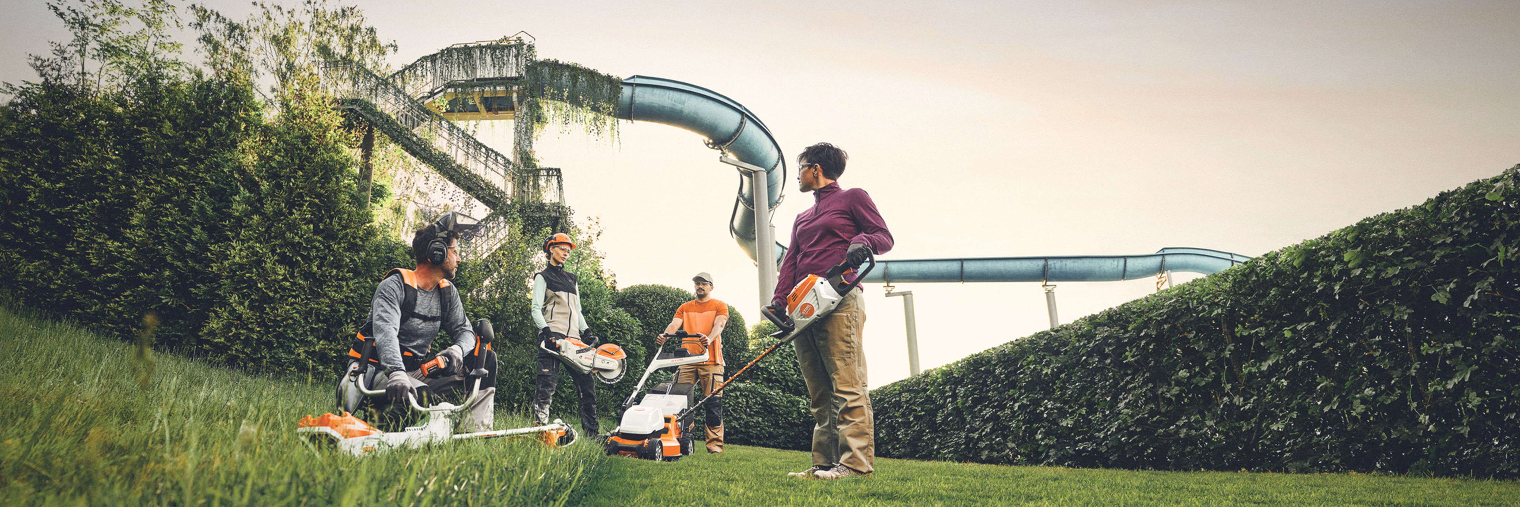Eine Gruppe von Personen arbeitet mit STIHL Gartengeräten in einer Grünanlage, mit einer großen Hecke und Wasserrutschen im Hintergrund. Sie sind mit Freischneidern, Rasenmähern und Schutzausrüstung ausgestattet.