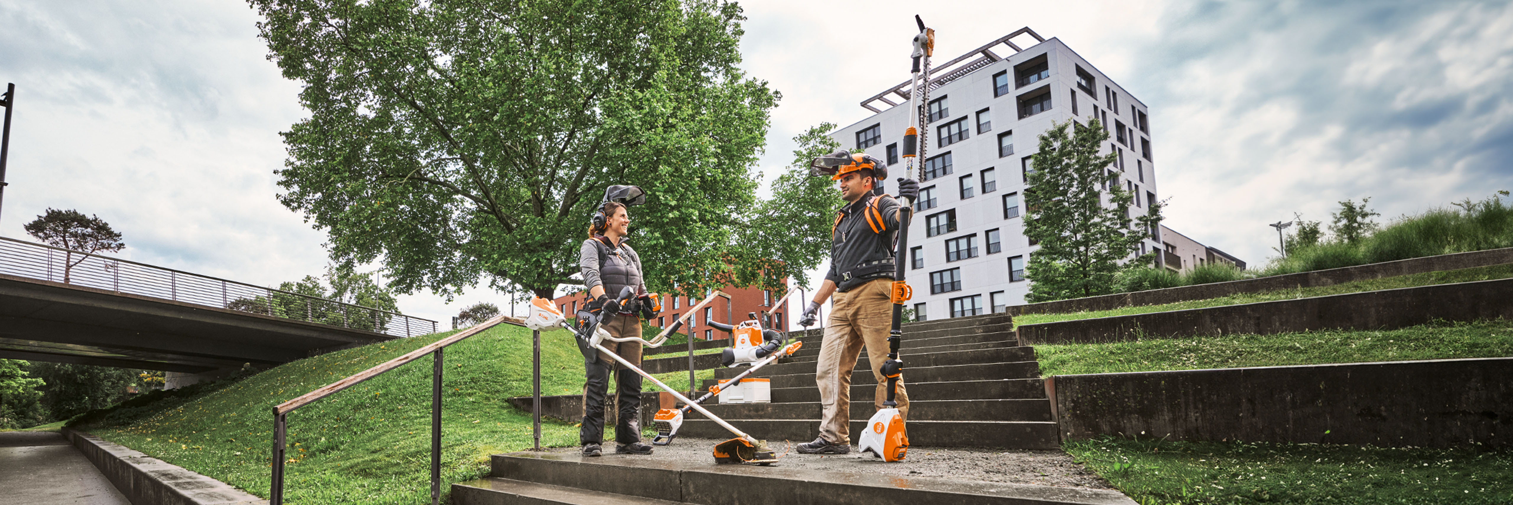 Zwei Landschaftsbau-Profis mit STIHL Pro-Geräten und Akkugeräten auf einer städtischen Grünfläche mit Treppen und Bäumen.
