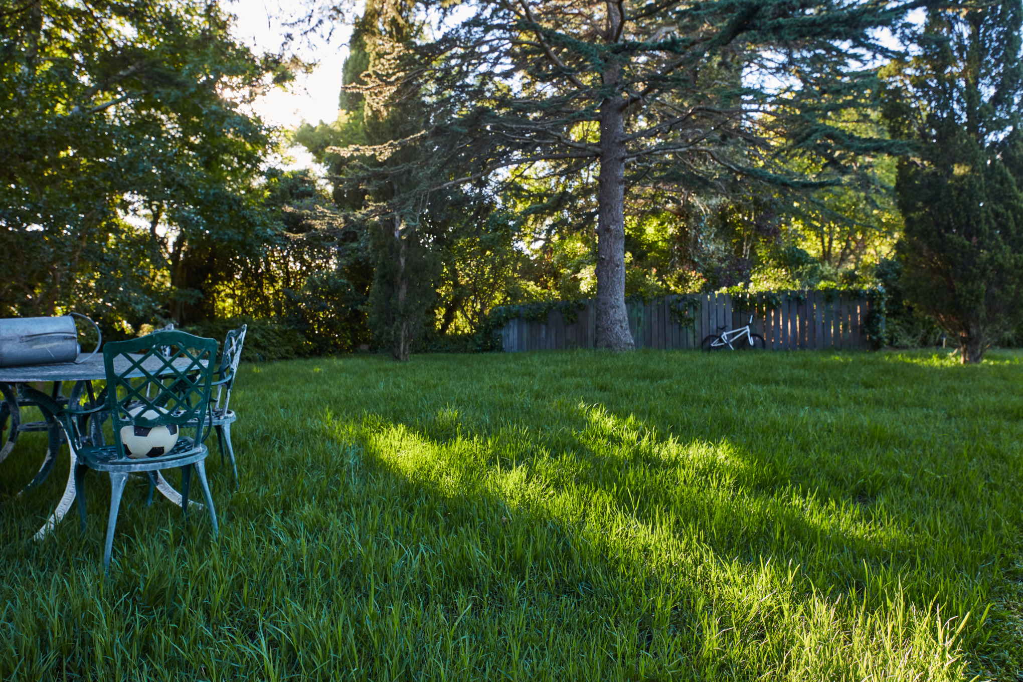 Schattenrasen mit Gartentisch und Stühlen, im Hintergrund lehnt ein Fahrrad an einem Holzzaun