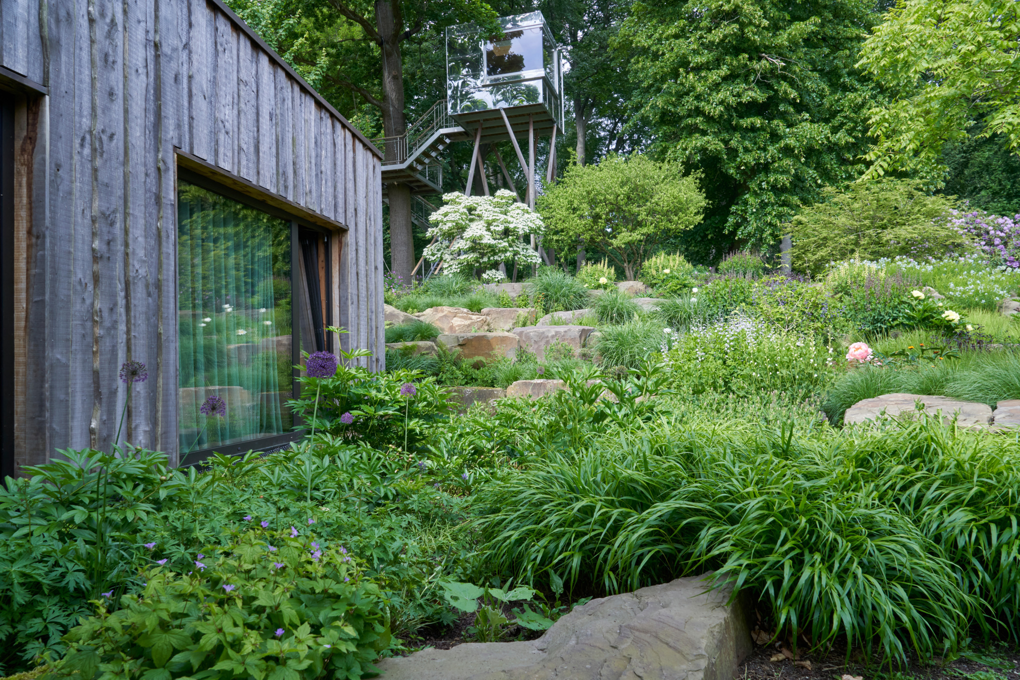Ein Hanggarten mit Steinen an einem Holzhaus, im Hintergrund ein Aussichtspunkt