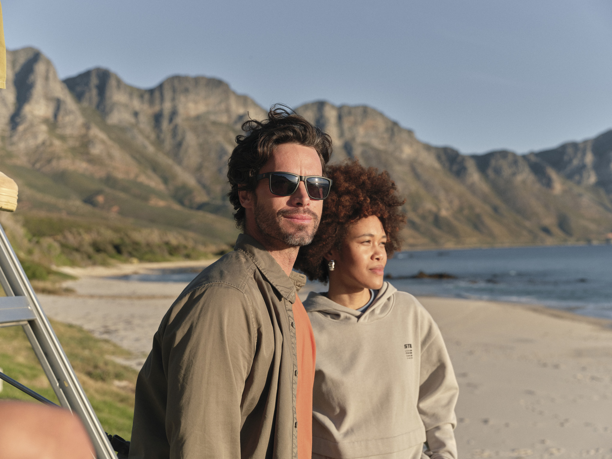 Mann und Frau, die Kleidung aus der STIHLcollection Fan Kogel Bay Kollektion tragen, stehen am Strand und blicken auf das Meer hinaus.