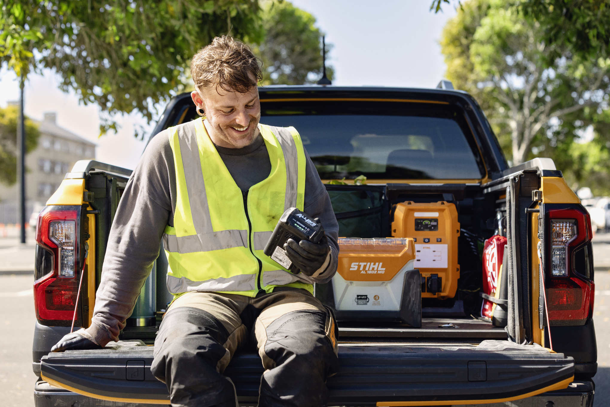 Mann sitzt auf einer Pickup-Laderampe und checkt den Ladestand seines ALLPRO-Akkus. Im Hintergrund sind das STIHL Schnellladegerät AL 1802 MO und der mobile Generater PS 3000