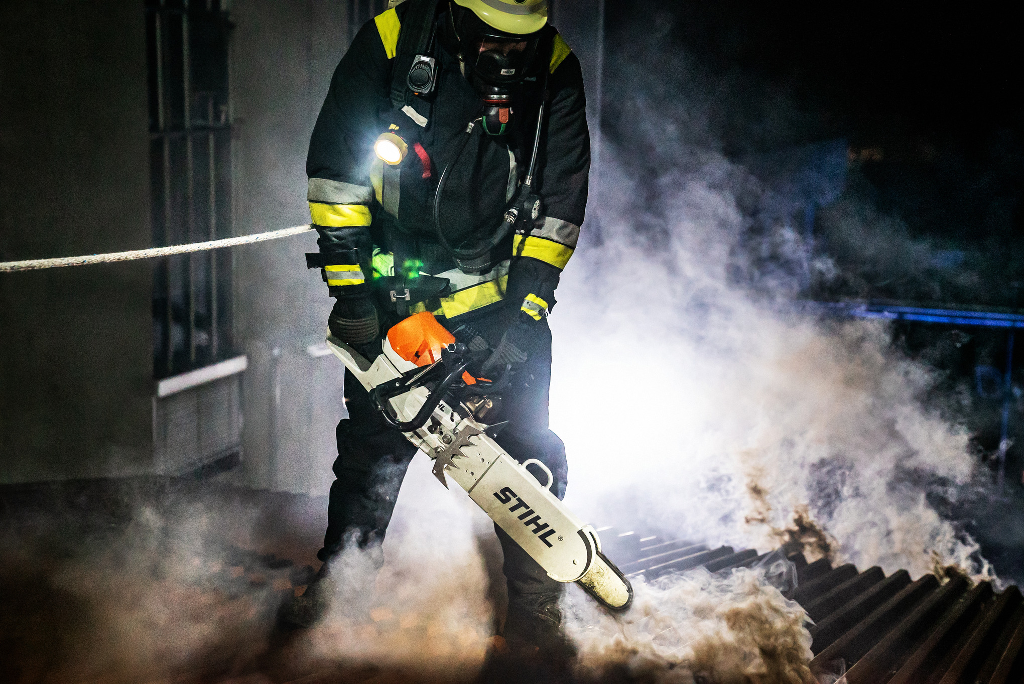 Person in Brandschutzausrüstung  mit STIHL Motorsäge auf einem brennenden Wellblechdach im Dunkeln 