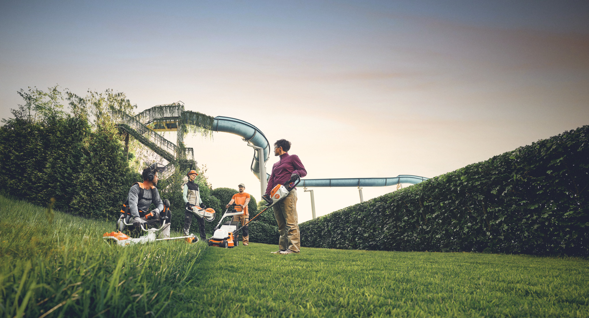 Eine Gruppe von Personen arbeitet mit STIHL Gartengeräten in einer Grünanlage, mit einer großen Hecke und Wasserrutschen im Hintergrund. Sie sind mit Freischneidern, Rasenmähern und Schutzausrüstung ausgestattet.