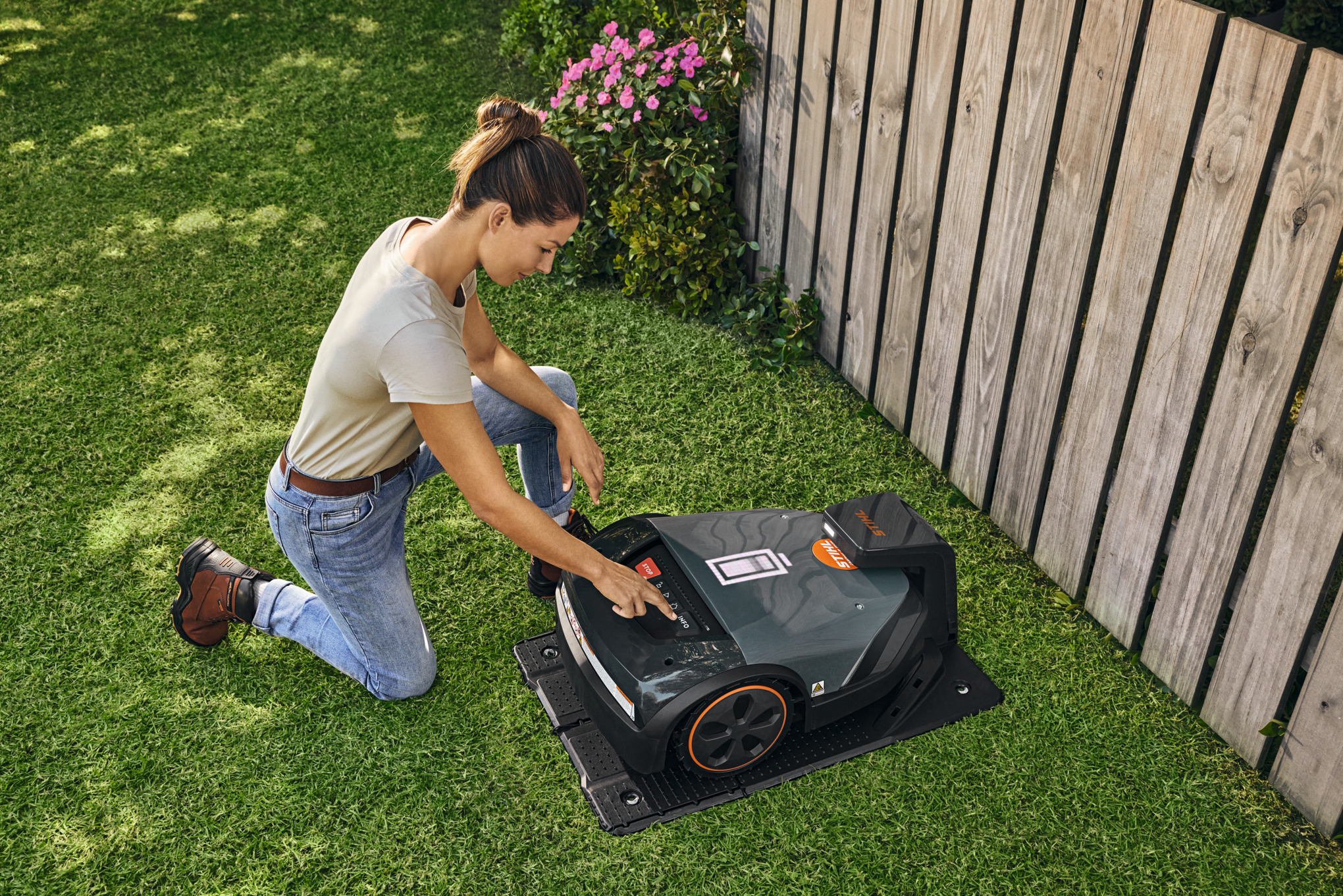 STIHL iMOW® Mähroboter in seiner Dockingstation im Garten während eine Frau etwas am Mähroboter einstellt.