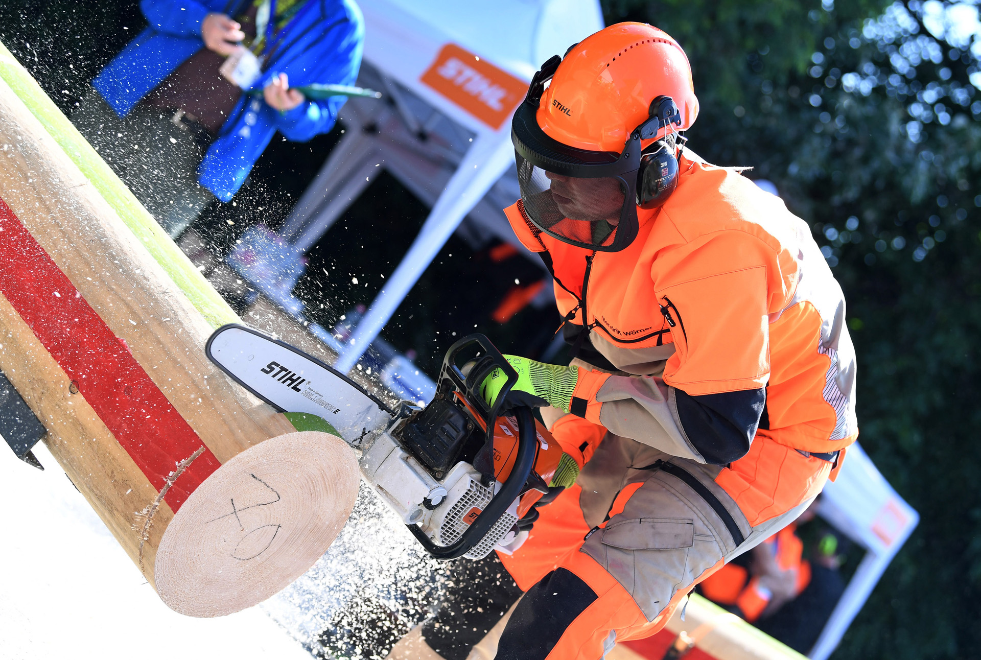 Mann mit STIHL Motorsäge zersägt einen Baumstamm, im Hintergrund ein Zelt mit dem STIHL Logo