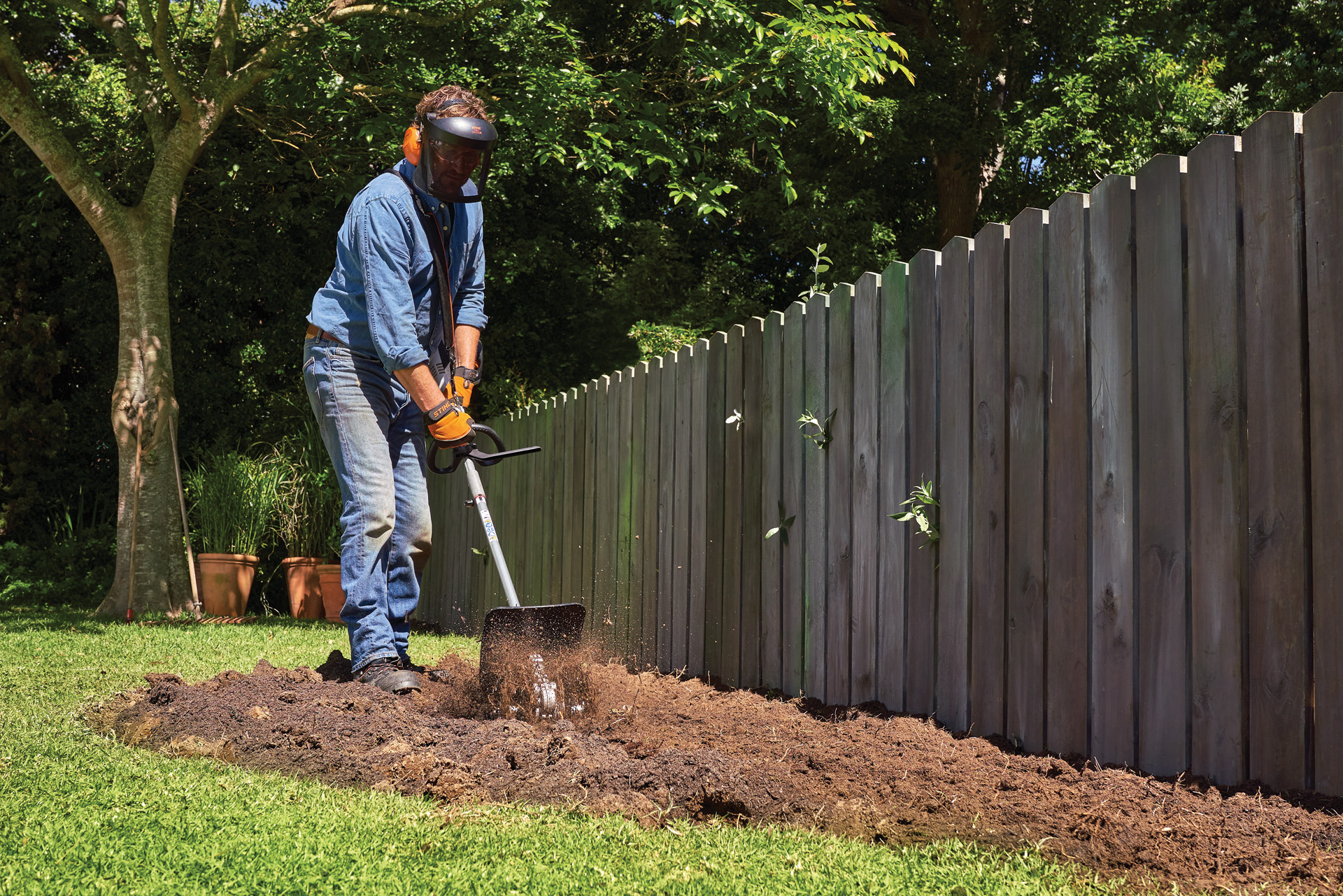 Person mit Gesichts- und Gehörschutz gräbt eine Fläche im Garten für ein Staudenbeet mit einem STIHL KombiSystem mit Bodenfräse um