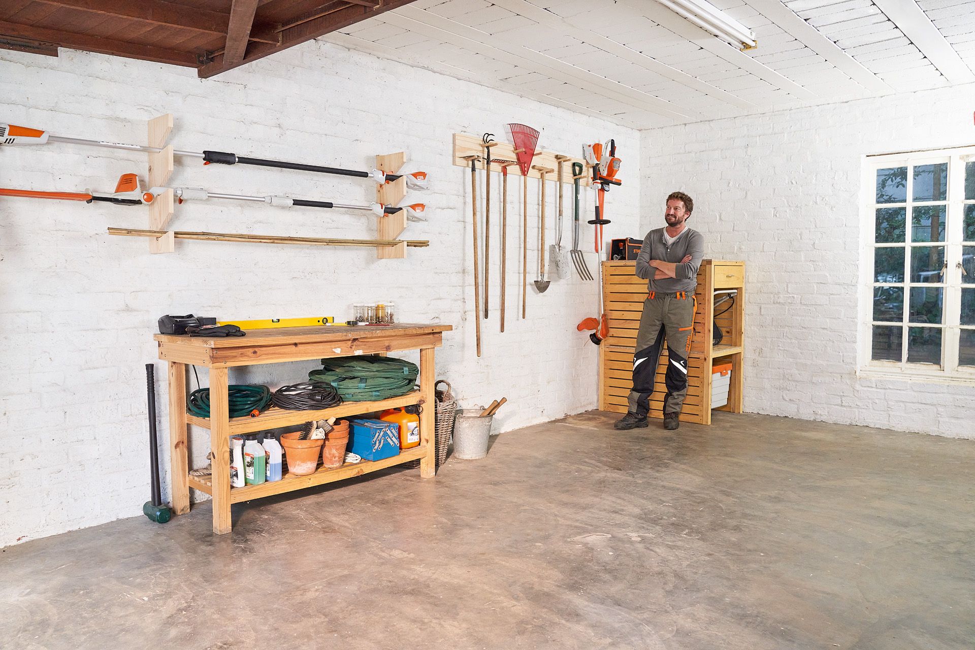 Ein Mann lehnt mit verschränkten Armen neben einem selber gemachten Gartengerätehalter in einer Garage 