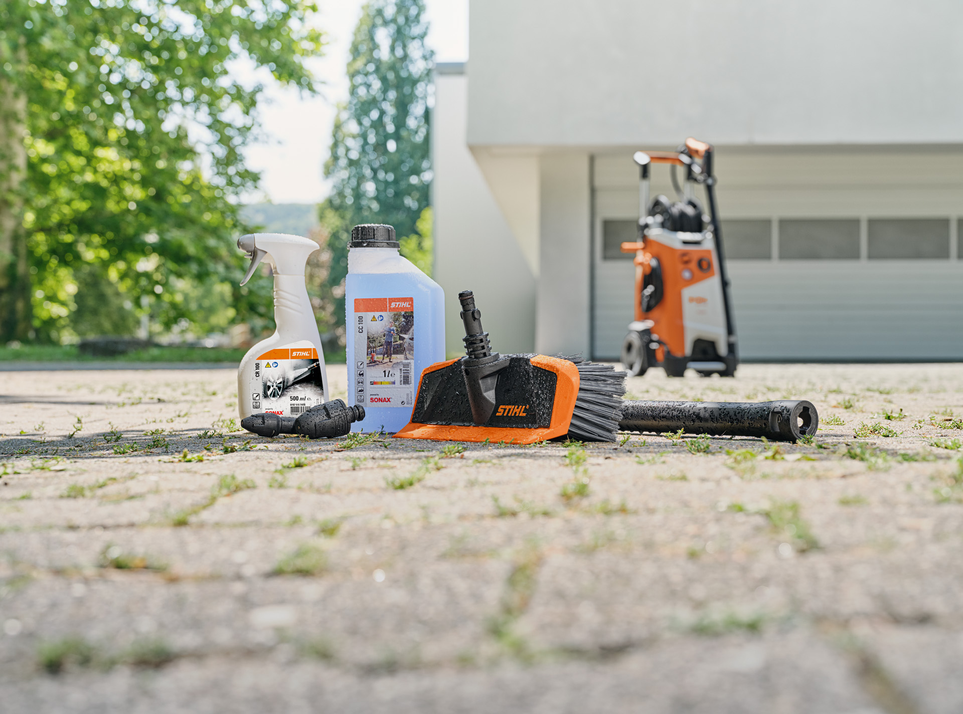  STIHL Felgenreiniger, Fahrzeugshampoo und Waschbürste auf dem Boden vor weißer Garage. Im Hintergrund ein STIHL Hochdruckreiniger