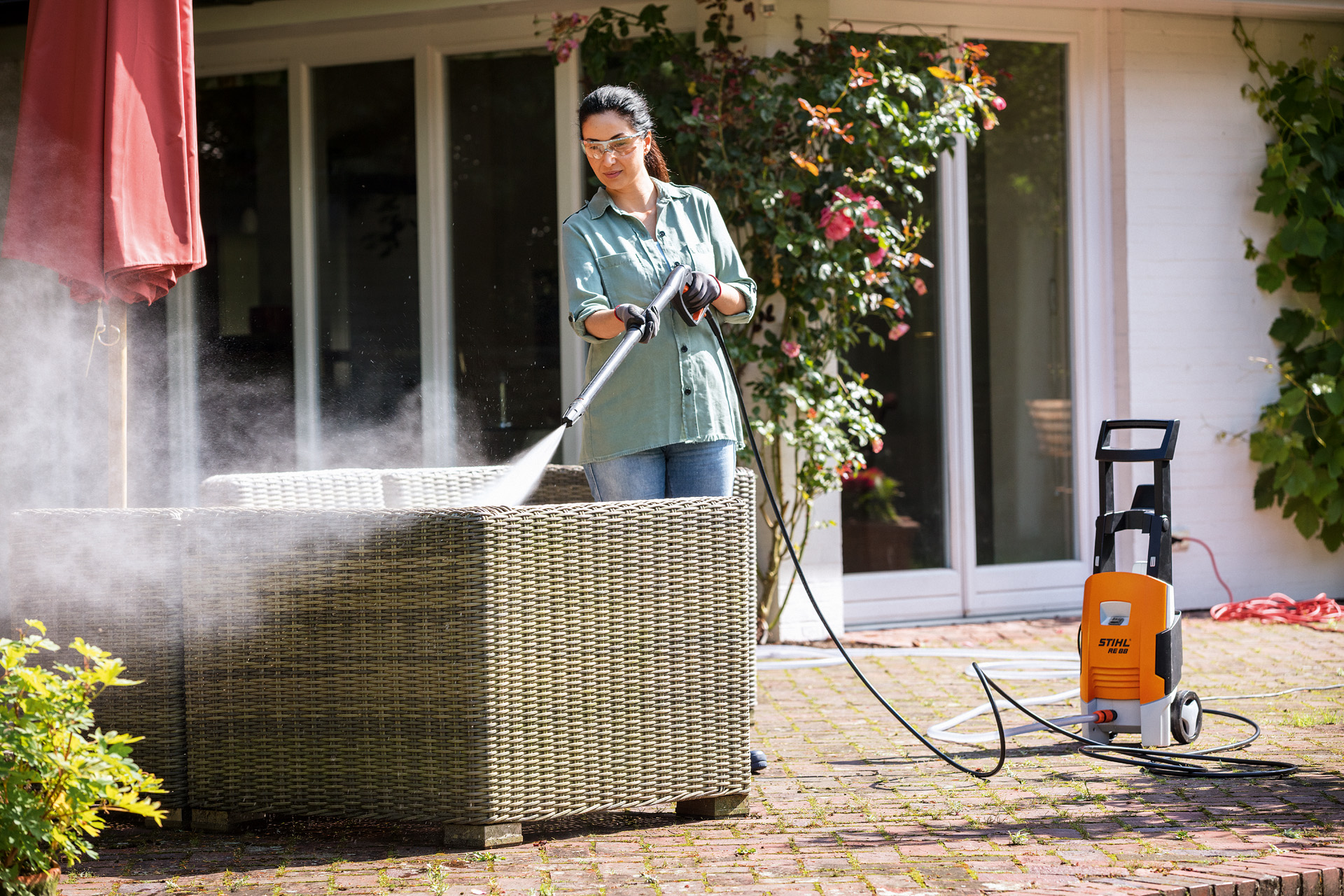 Frau mit Schutzbrille ist dabei, mit dem STIHL Hochdruckreiniger RE 88 auf der Terrasse ihre Gartenmöbel zu reinigen