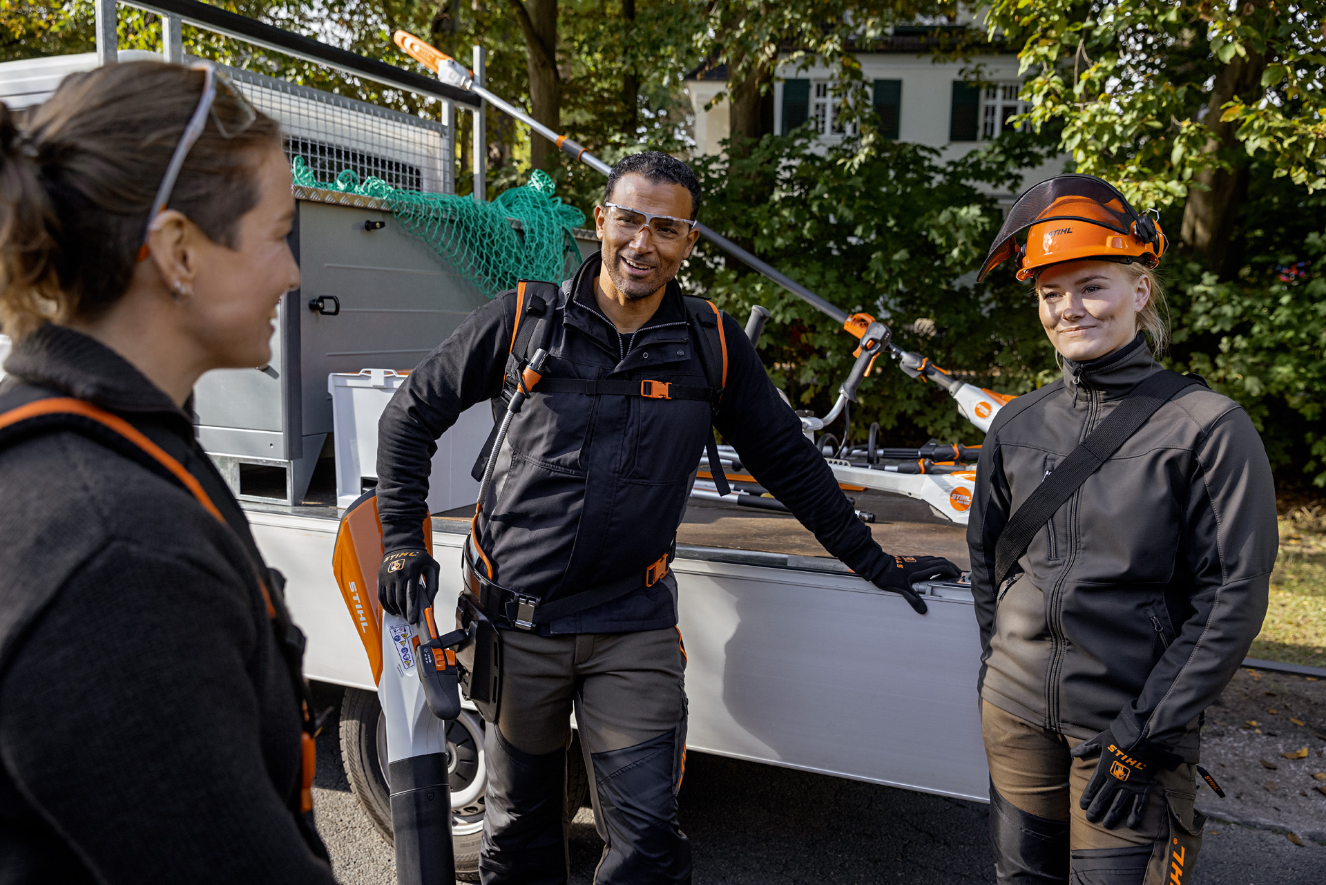 Drei Personen in STIHL Ausrüstung vor einem Fahrzeug, teilweise mir Gartengeräten 