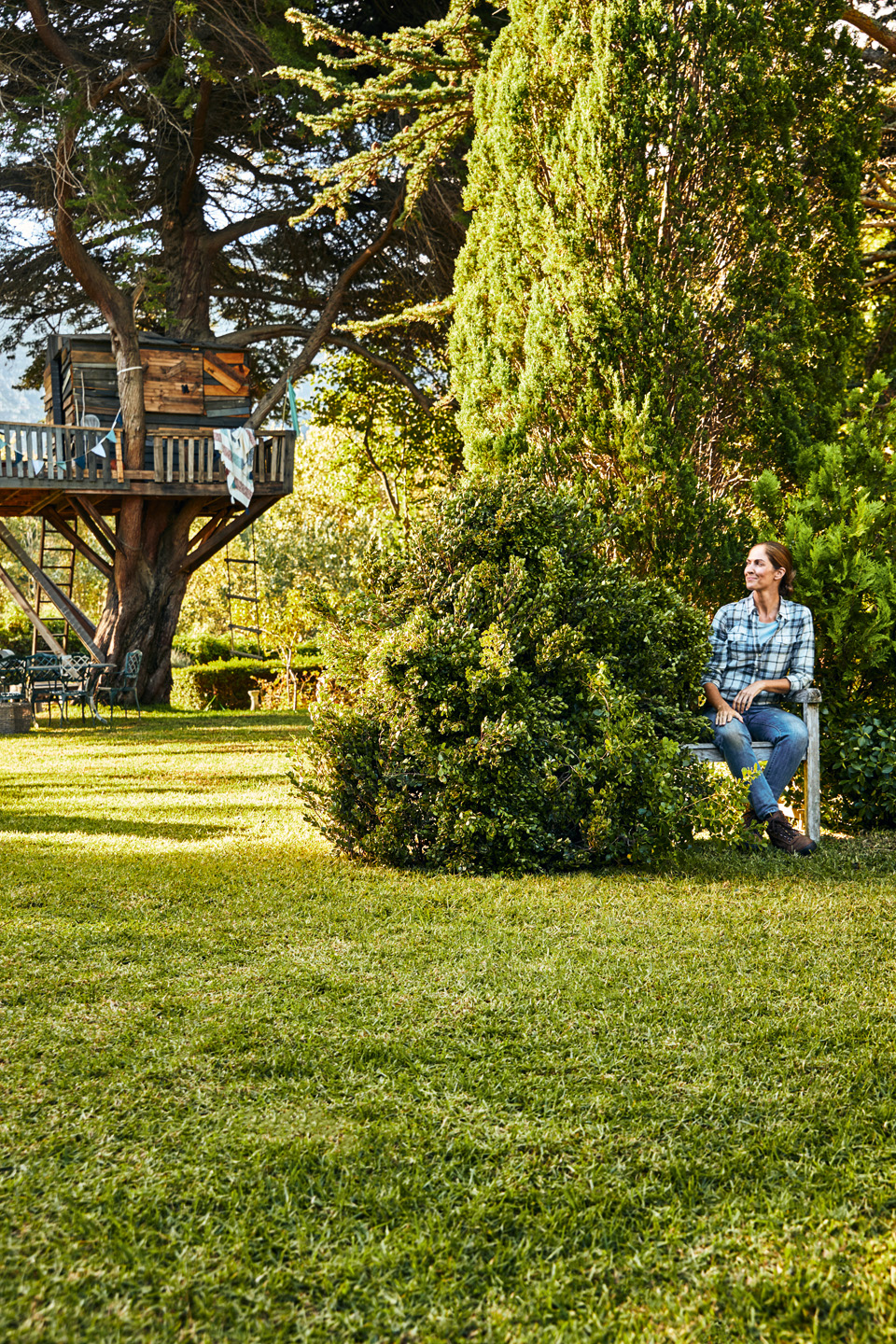 Frau in karierter Bluse sitzt auf einer Gartenbank hinter einem Busch auf einem grünen Grundstück mit Baumhaus im Hintergrund