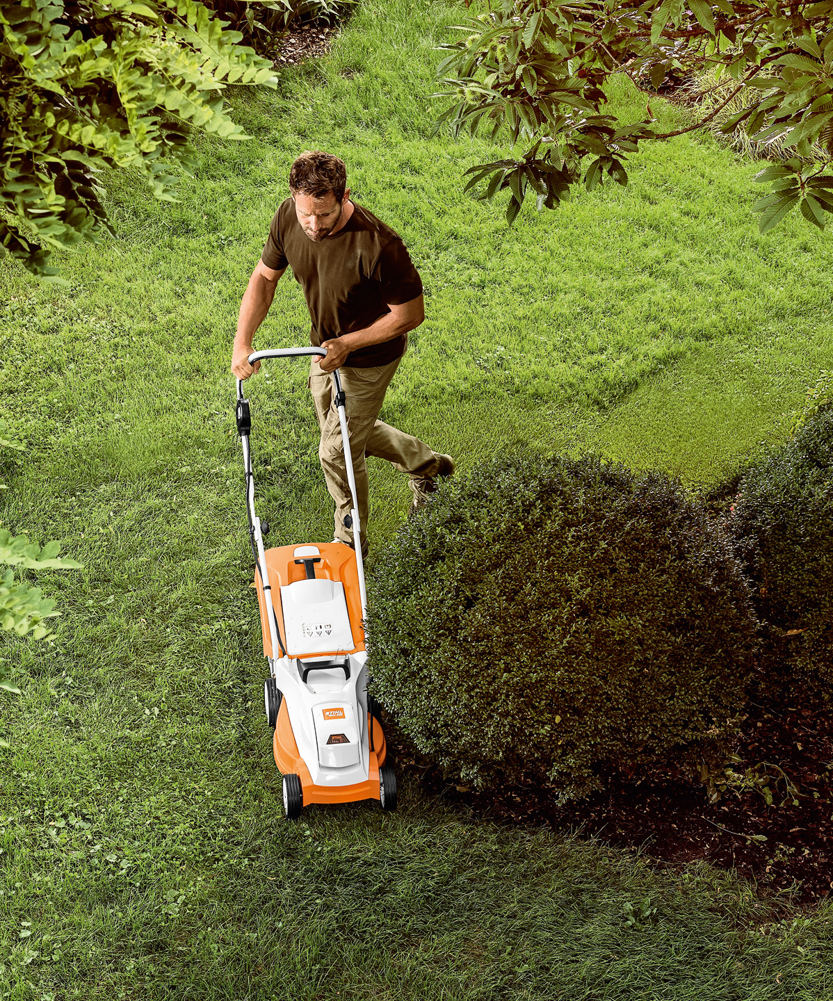 Blick von oben: Mann mäht eine Rasenfläche mit STIHL Akku-Rasenmäher RMA 235 rund um kleinen Baum herum.