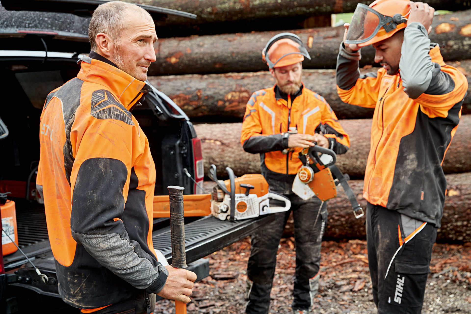 Drei Waldarbeiter in persönlicher Schutzausrüstung vor einem Holzscheit im Wald.