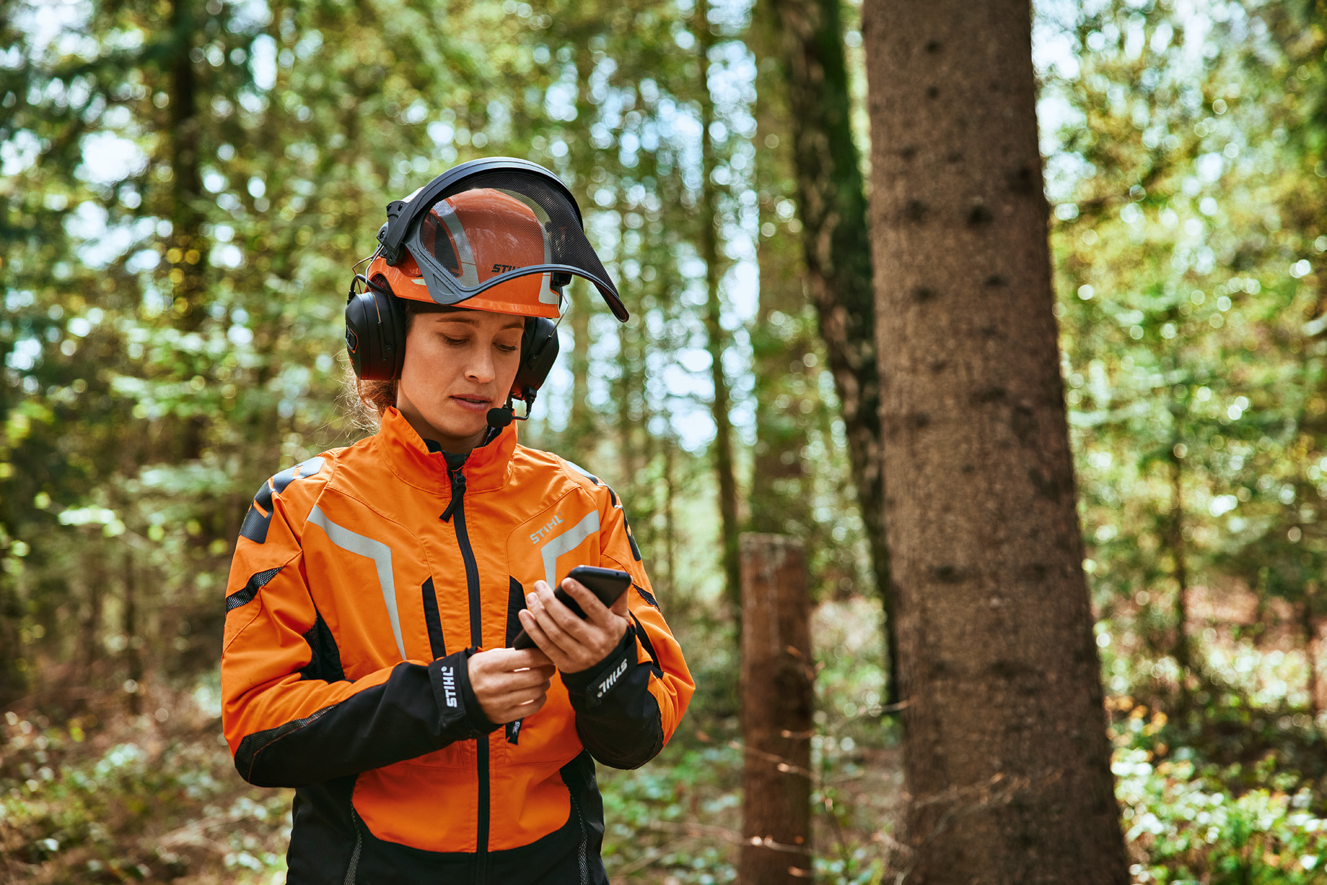 Eine Forstarbeiterin steht in persönlicher Schutzausrüstung von STIHL inklusive dem Helmset ADVANCE ProCOM in einem Wald und nutzt die STIHL Holzliste App.