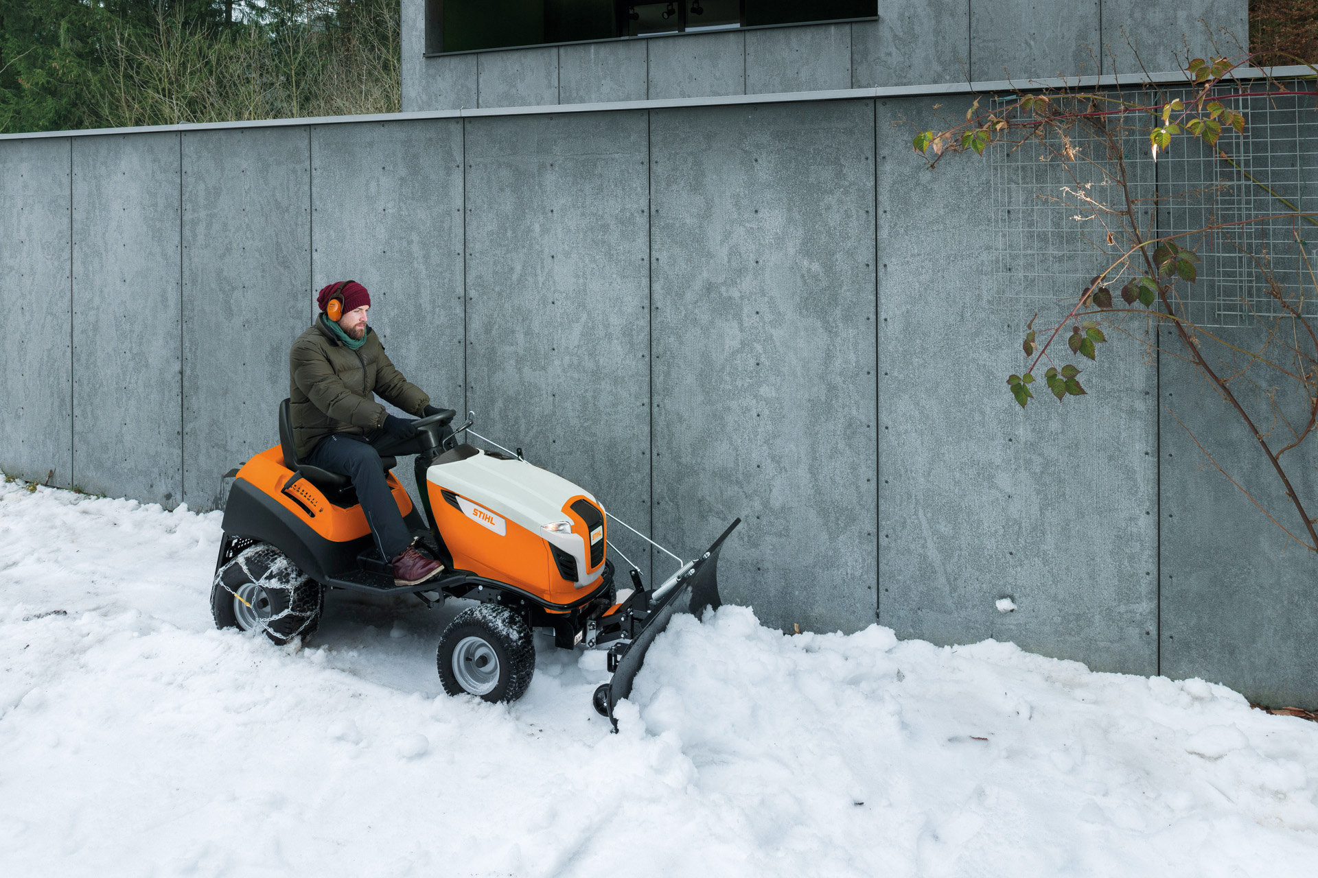 Mann räumt Schnee vor einer Mauer auf einem Rasentraktor mit Schneeschild 