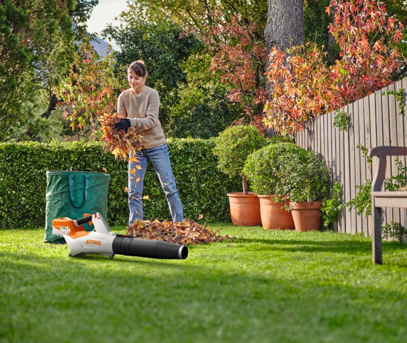 Eine Frau räumt mithilfe eines Akku-Laubbläsers STIHL BGA 60 Laub in einem Garten auf