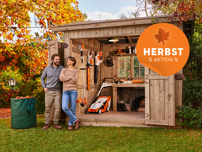 Eine Frau und ein Mann stehen in ihrem herbstlichen Garten vor ihrem Geräteschuppen.