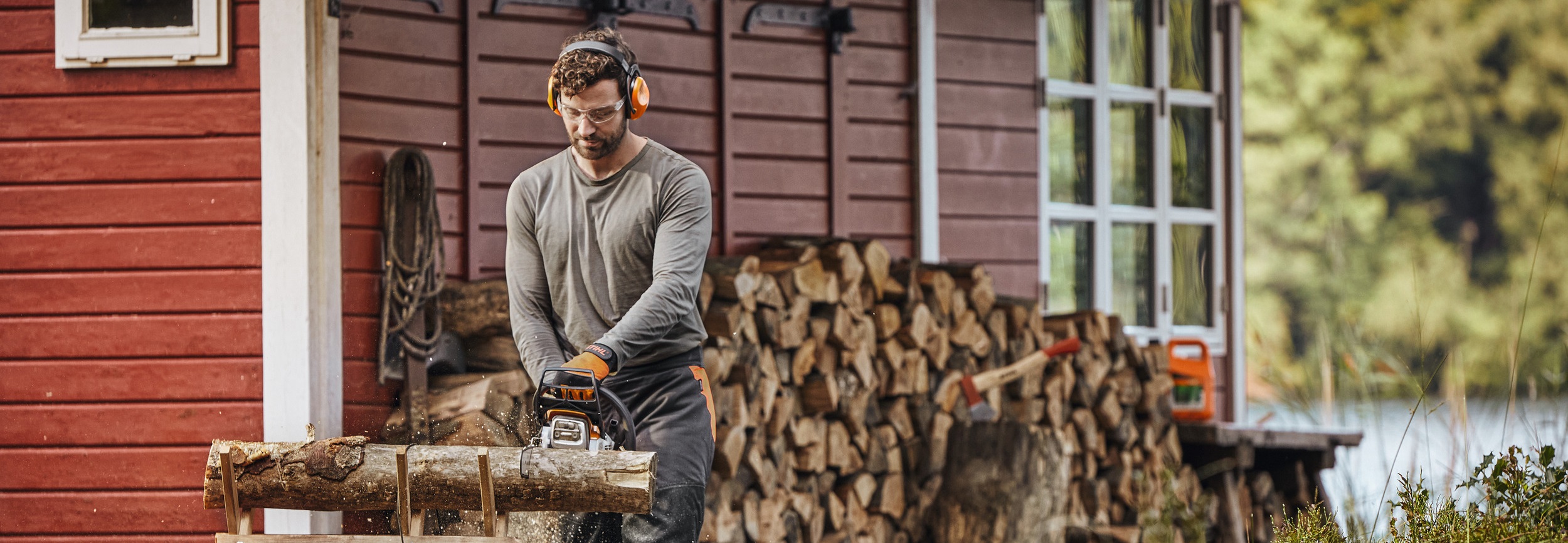Ein Mann mit STIHL Akku-Motorsäge sägt einen Holzstamm, im Hintergrund ist eine rote Holzhütte zu sehen.