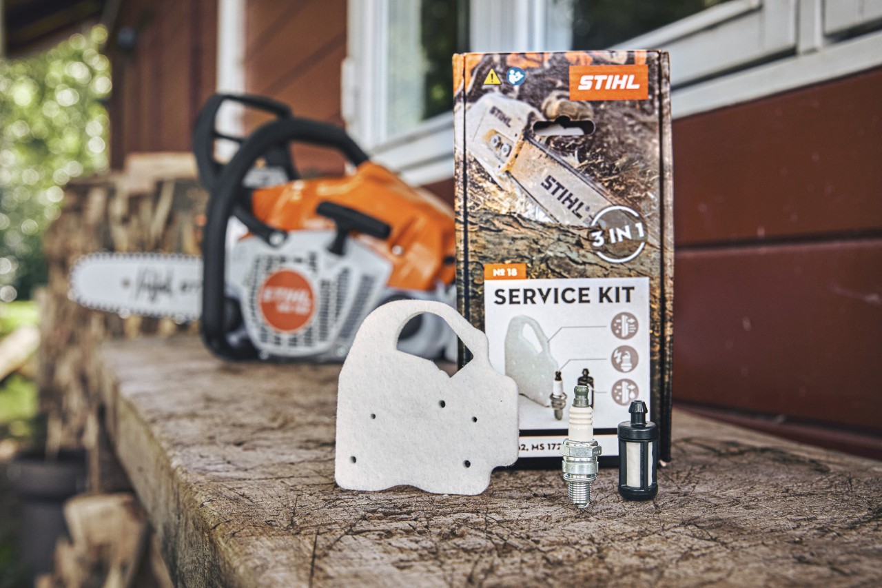 STIHL Service Kit 18 auf einer Werkbank, davor die einzelnen Komponenten Vliesluftfilter, Zündkerze und Kraftstofffilter