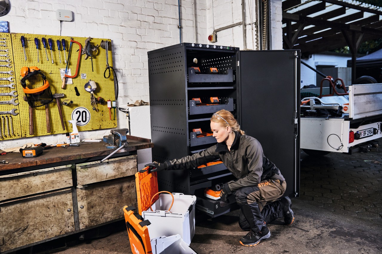 Blonde Frau in STIHL Arbeitskleidung in einer Werkstatt mit Akku-Ladeschrank und STIHL connected Box, im Hintergrund eine Werkzeugwand 
